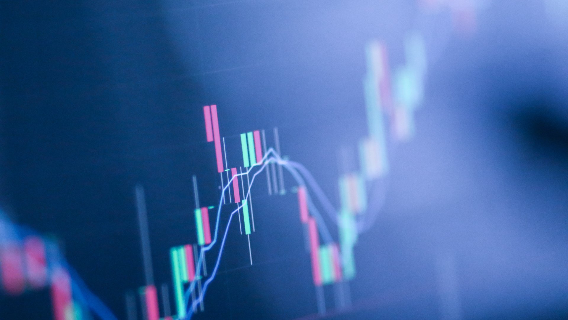 Close-up of a computer screen displaying a blurred candlestick stock chart in pink and teal against a dark navy-blue background, with an out-of-focus blue trend line.