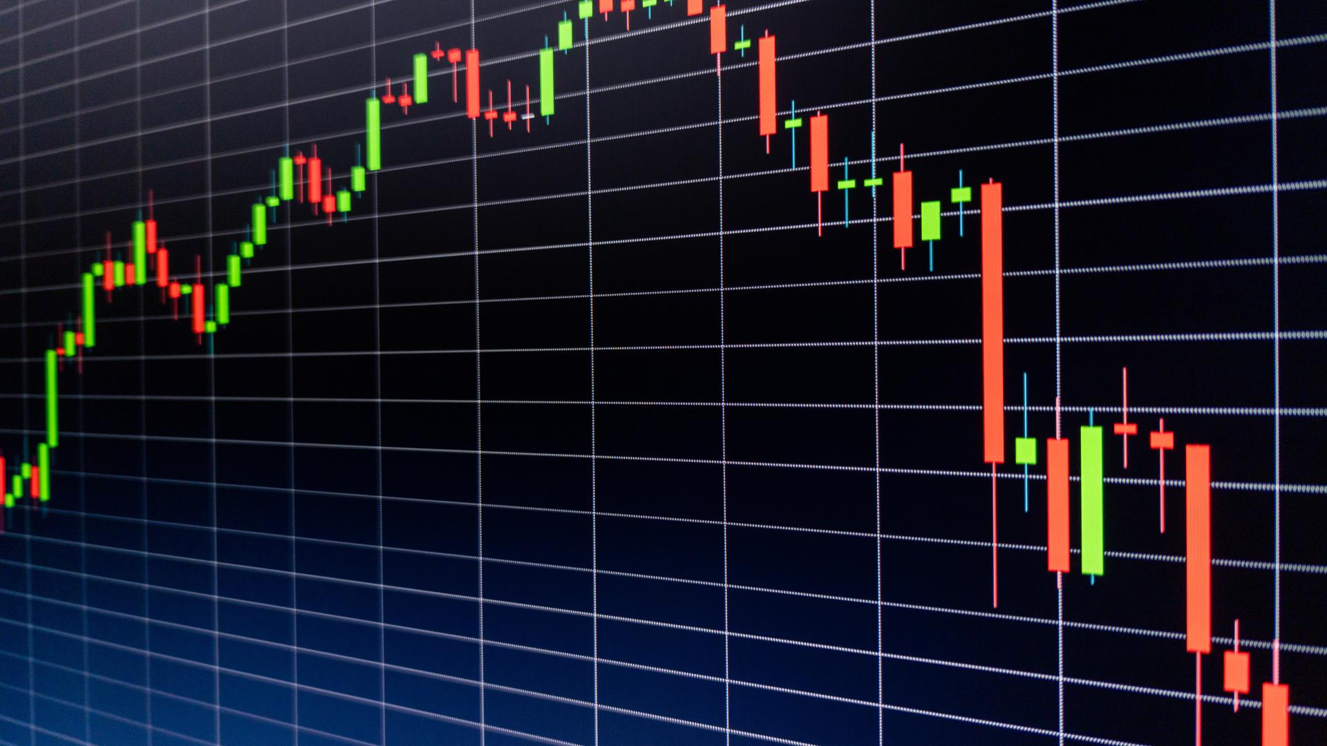 Dark trading screen viewed at an angle, showing a candlestick price chart in green and red climbing then reversing into a steep decline against a navy-black grid.